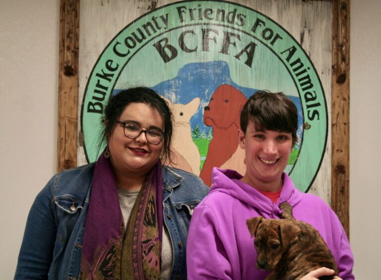 Two people smiling with a brindle puppy in front of the shelter sign